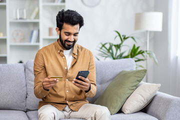 Young joyful man sitting on sofa at home, using application on phone for online shopping in online...