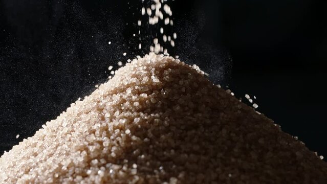 Brown cane sugar is poured into pile. Close up slow motion black background. Prudent control of sugar intake helps mitigate risk of obesity, diabetes, and other diseases that arise from it. Nutrition
