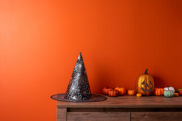 Witch hat and pumpkins on  wooden chest of drawers opposite  orange wall. Space for text. Greeting...