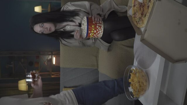 Girl watches movie and eats popcorn. Boy comes and brings glasses with whiskey, she declines looks negative. Vertical video. HDR BT2020 HLG Material.