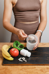 Athletic woman in sportswear with measuring spoon in her hand puts portion of whey protein powder into a shaker on wooden table with amino acid white capsules, bananas and apple, making protein drink