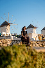 Young woman in black dress at the windmills in Mykonos - obrazy, fototapety, plakaty