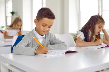 Little focused boy draws in workbook during studies completes assignment from teacher wants to get good grade sits at desk in elementary school. Quality education, back to school concept