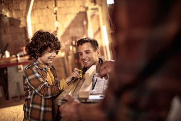 Young father teaching his son how to measure wood in a carpentry shop