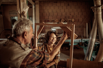 Senior male carpenter showing his grandson a wood frame in a carpentry shop