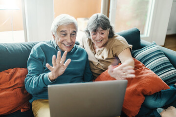 Senior couple on sofa using laptop for video call, showcasing elderly tech adaptation, online family connection, remote communication, digital lifestyle.