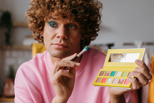 Confident young gay man holding eyeshadow palette while presenting make-up products from home