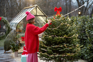 Young woman in red decorates lush Christmas tree with festive ballls and garland at backyard of her...