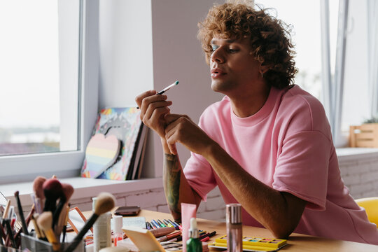 Confident young gay man blowing on eyeshadow brush while applying make-up at home - Powered by Adobe