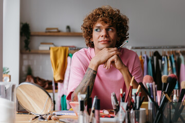 Dreamful young gay man looking away while applying make-up at home