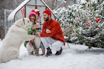 Man and woman play with their dog, while spending happy winter time together at snowy backyard with Christmas tree on background. Young family celebrate winter holidays
