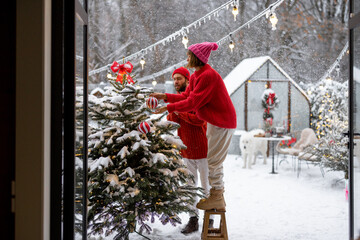 Man and woman decorate Christmas tree with festive balls, while preparing for a winter holidays at snowy backyard of their house. Happy family celebrating New Year's holidays