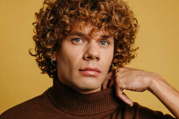 Fashionable curly young man adjusting sweater neck and looking at camera against yellow background