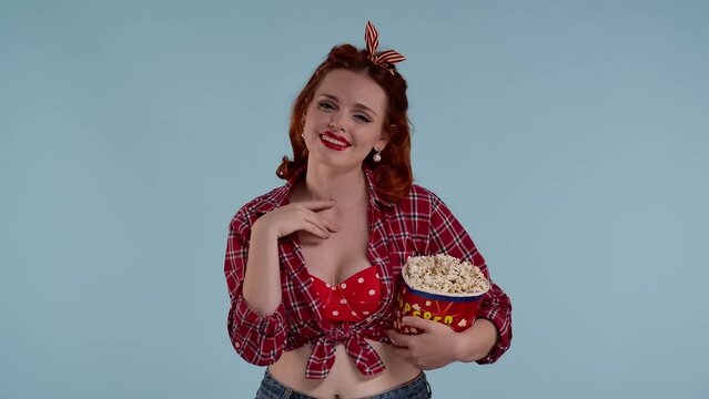 In the frame on a blue background, a young red-haired woman with bright makeup. Demonstrates watching a movie in a theater. She is holding popcorn and laughing she likes it she is joyful, amused