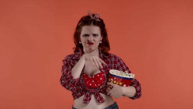 In the frame on a red background is a young red-haired woman with bright makeup. She demonstrates watching a movie. She does not like it she is not happy and throws popcorn. HDR BT2020 HLG Material.