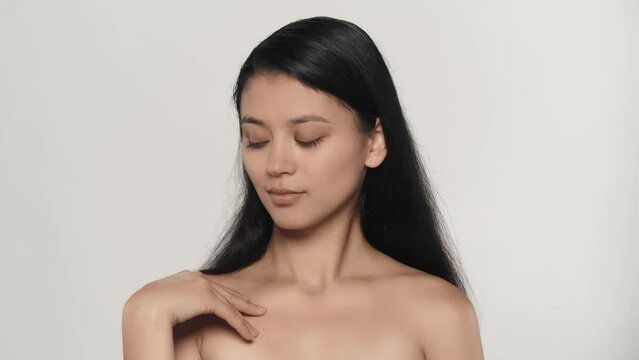 Closeup studio shot of beautiful female model. Brunette girl with long hair and natural makeup touching collarbone area smooth skin and smiles at the camera.