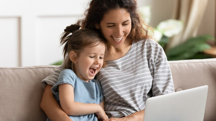 Overjoyed young mother and funny little daughter sit rest on sofa at home laugh watch funny video on laptop, happy millennial mom and small girl child relax on couch have fun using computer together