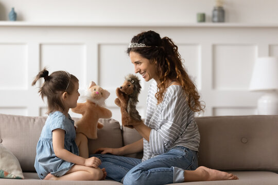 Happy young mother relax on couch in living room play puppet theater with cute preschooler daughter, smiling millennial mom or nanny have fun enjoy childish game rest with little girl child at home