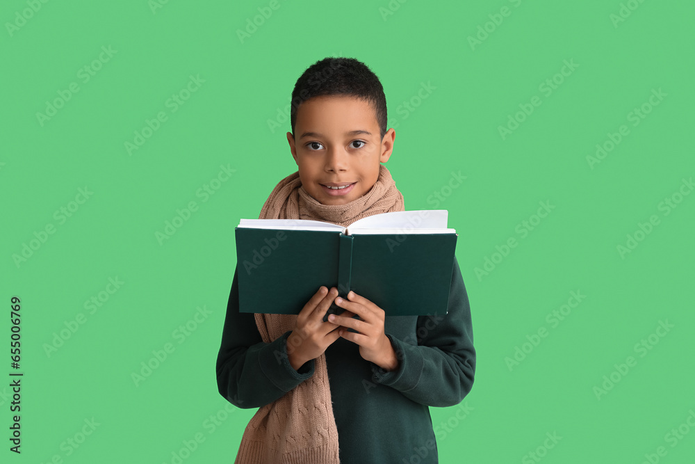 Poster Cute African-American boy in winter clothes reading book on green background