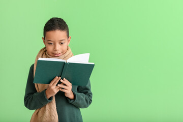 Cute African-American boy in winter clothes reading book on green background