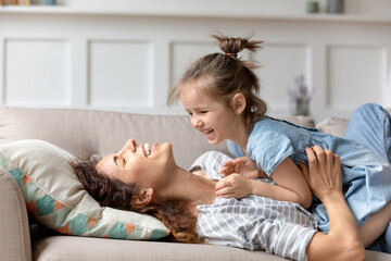Overjoyed millennial mom rest on couch in living room playing with happy preschooler daughter,...