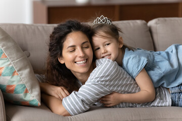 Happy small preschooler girl lying with excited young mother hugging and cuddling on couch at home, cute little daughter embrace smiling millennial mom relax together on sofa, show love and affection