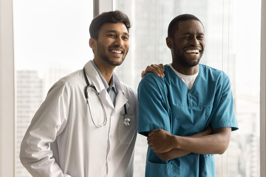 Happy multiethnic young medical colleagues men, cheerful Indian practitioner and African surgeon standing together, looking at camera away, laughing, having fun. Healthcare jobs friends portrait