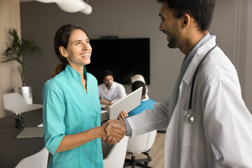 Clinic staff boss, chief doctor giving handshake to happy surgeon with respect, recognition, gratitude. Positive medical colleagues shaking hands on corporate meeting, smiling, laughing - obrazy, fototapety, plakaty