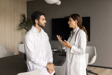 Fototapeta premium Two serious male and female doctor colleagues in white coats talking in meeting room, discussing medical practice, clinic work process, treatments cases, physician occupation, job