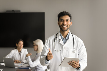 Fototapeta premium Happy young Indian doctor in white coat showing like hand, making thumb up gesture at camera, holding digital tablet computer, posing with colleagues meeting and talking in background
