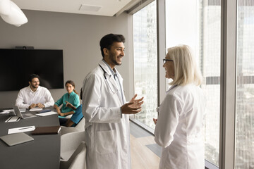 Obraz premium Positive Indian younger doctor man and elder colleague woman talking in meeting room, smiling, laughing, discussing medical practice, enjoying networking, job communication