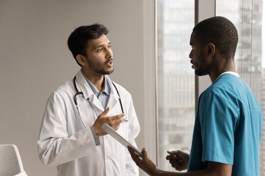 Multiethnic practitioner and surgeon men discussing diagnosis, medical examination result, using tablet computer, talking in clinic, hospital office. Indian doctor instructing nurse