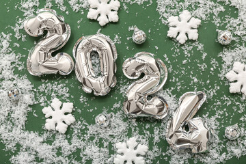 Composition with figure 2024 made of foil balloons, snow and Christmas decorations on green background