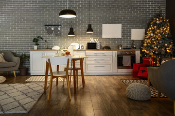 Interior of modern kitchen with Christmas tree, glowing lamps and dining table at evening