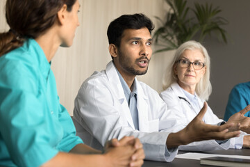 Serious young Indian physician man consulting colleagues about difficult case, speaking at hospital staff meeting, council. Diverse team of doctors discussing medical problems