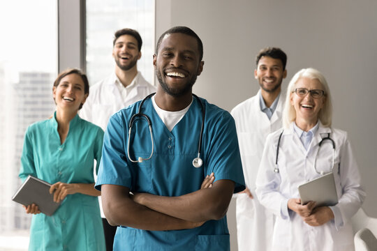 Happy African American surgeon man with folded arms standing in front of diverse colleagues, looking at camera, smiling, laughing. Small clinic head doctor, medical business leader portrait