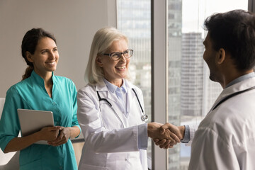 Happy senior clinic boss woman shaking hands with young male doctor, smiling, laughing, hiring new...