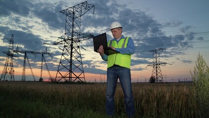 Worker conducts examination of power transmission lines with laptop at sunset