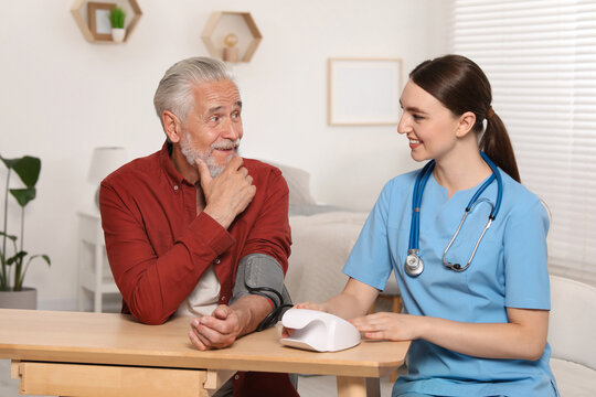 Young healthcare worker measuring senior man's blood pressure at wooden table indoors