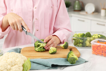 Woman cutting green broccoli near containers with fresh products at white marble table in kitchen, closeup