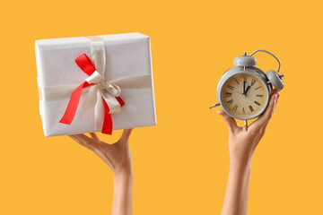 Hands holding Christmas gift and alarm clock on yellow background