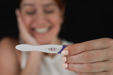 Blurred portrait of happy caucasian woman holding positive express pregnancy test on black background. 