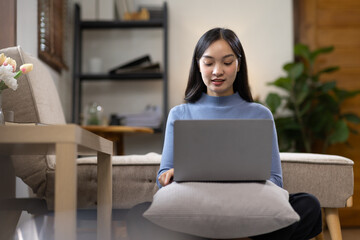 Woman sitting on floor reading book with laptop analyzing marketing or financial data online, calling watching elearning webinar. Happy asian female student studying at home.