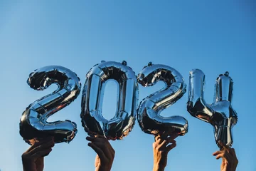 Rolgordijnen Silver foil number 2024 celebration new year balloon on blue sky background. Happy New year greetings concept. Hands holding balloons two thousand twenty-fourth year Christmas holiday concept  © anna.stasiia