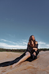 Fototapeta na wymiar A brunette in a black dress with a long train made of silk fabric sits on a sandy dune in the wind. Landscape of sand dunes and blue sky with clouds.