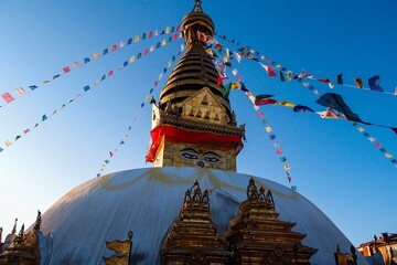The iconic main stupa of Swayambhunath, bathed in the warm hues of a Kathmandu sunset, stands tall against the sky, adorned with vibrant Tibetan prayer flags that dance gracefully in the breeze.
