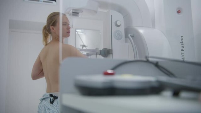 Adult woman stands topless during mammography screening checkup in hospital radiology room. Doctor presses button on control panel to launch mammogram machine using computer. Breast cancer prevention.
