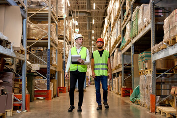 Retail Warehouse full of Shelves with Goods, Two Young Caucasian Male Workers Supervisors in Working Uniform and Helmet Discuss Product Delivery. Men Walking and Having Talk In Warehouse - obrazy, fototapety, plakaty