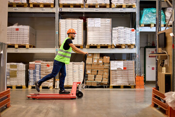 cheerful caucasian male staff dressed in working clothes and helmet in warehouse having fun standing on pallet truck jack, driving among shelves, enjoy free time, leisure at work place - obrazy, fototapety, plakaty
