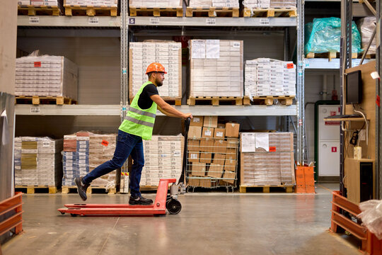 cheerful caucasian male staff dressed in working clothes and helmet in warehouse having fun standing on pallet truck jack, driving among shelves, enjoy free time, leisure at work place